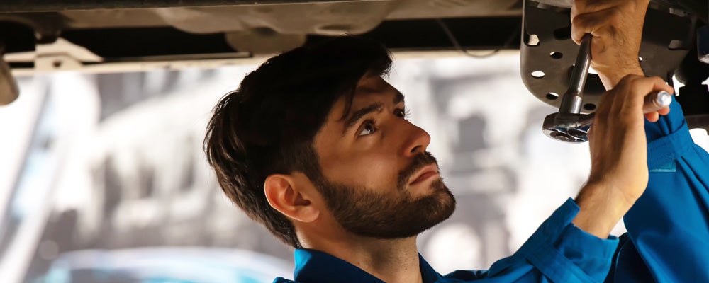 Close-up of a vehicle technician in a blue work uniform using a tool to repair or inspect the underside of a car