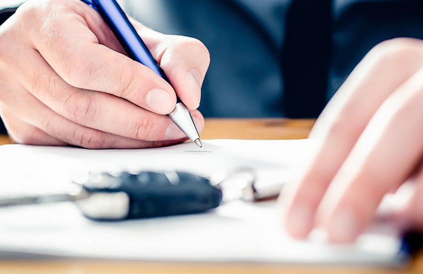 A hand is holding a blue-and-silver pen and writing or signing on a white sheet of paper.